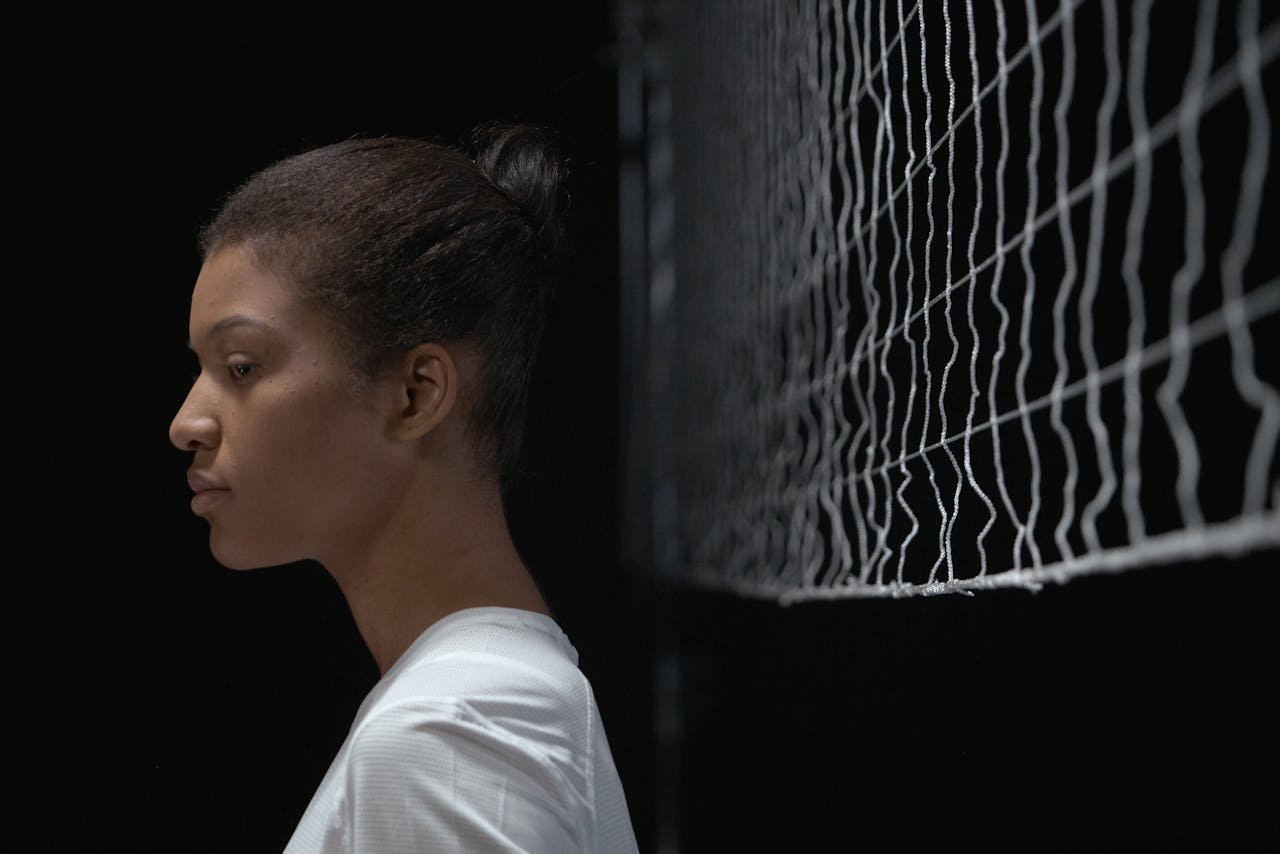 Young woman stands in profile beside a volleyball net, focused and serious.